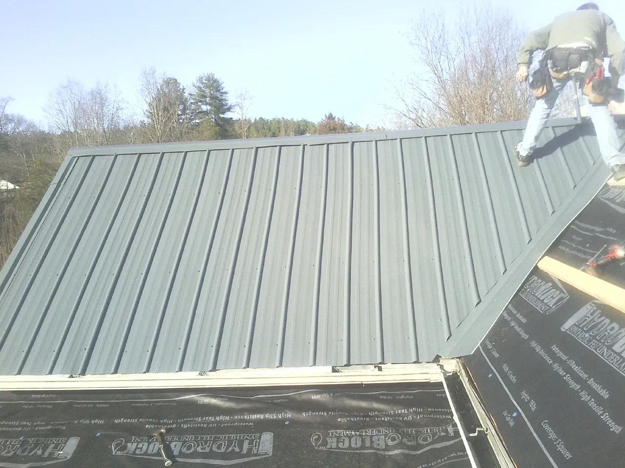 Crew installing dark green standing seam panels over synthetic underlayment in Townley