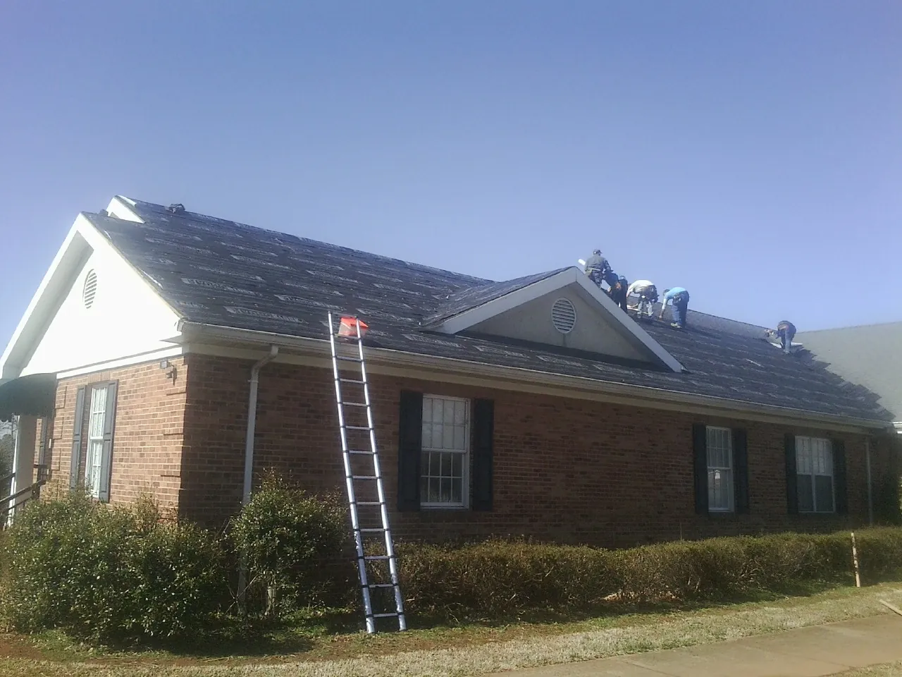 Crew tearing shingles off a brick ranch roof with a ladder set — Metal Roof Repair work by our crew in Townley