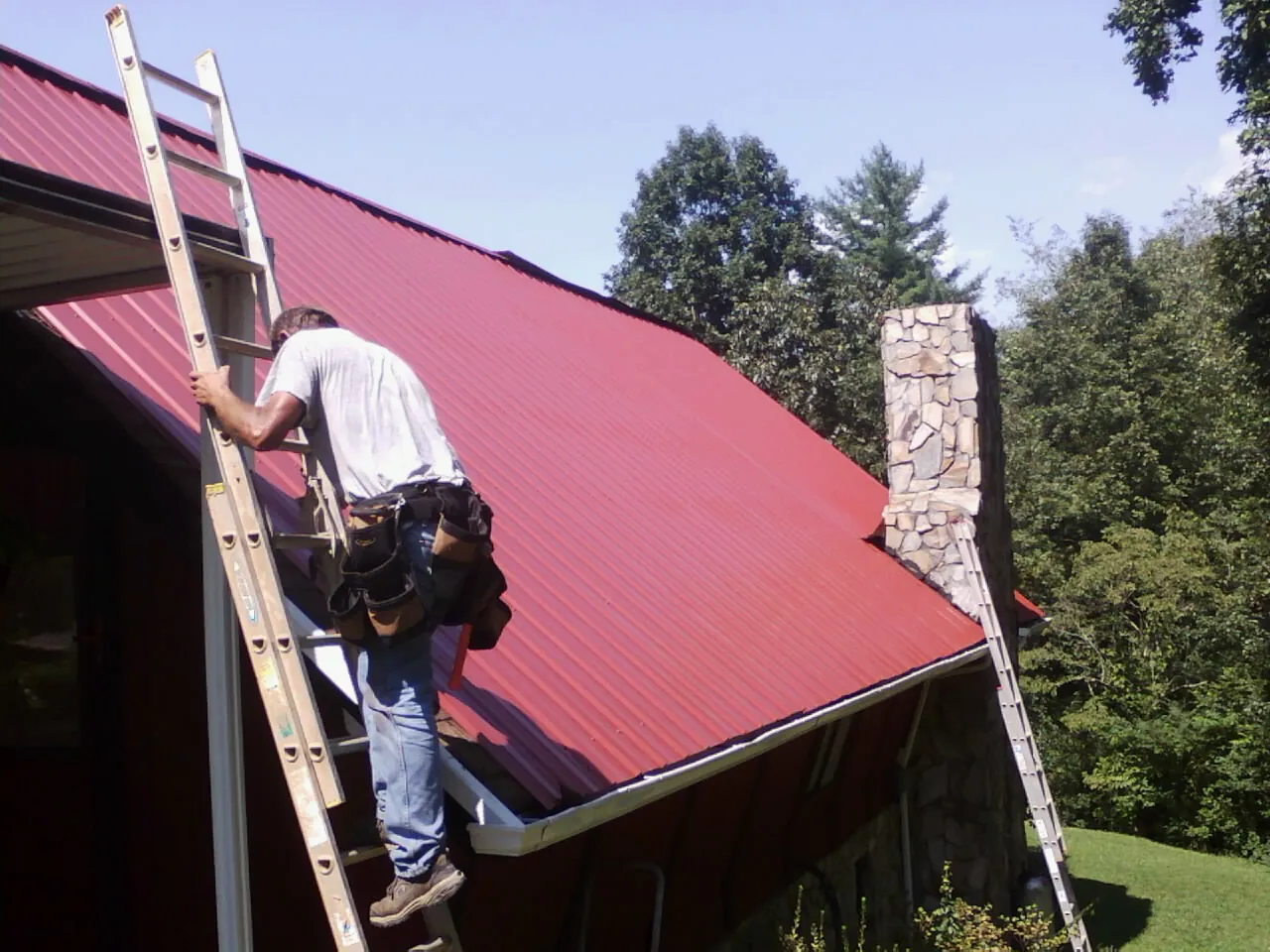 Roofer on a ladder beside a red metal roof and stone chimney — Soffit & Fascia Repair by Roofing Brothers in Townley